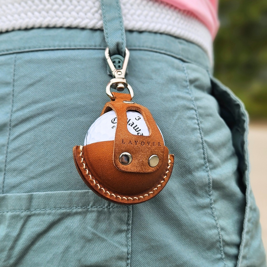 Brown leather golf ball holder attached to a belt, worn on pants, with a golf ball securely placed inside. The holder features a smooth leather texture, precise stitching, and a compact design for convenient access