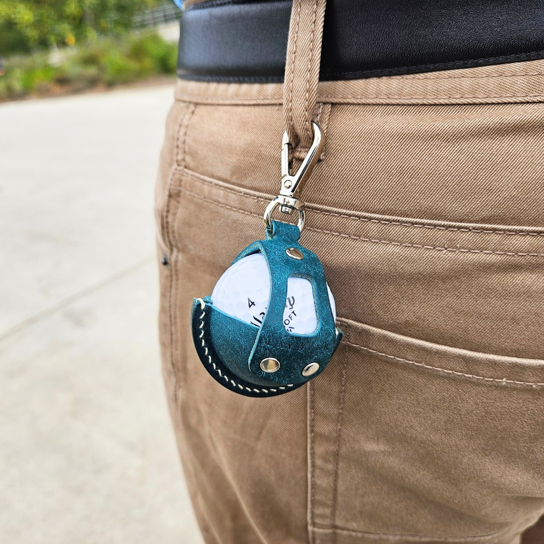 Green leather golf ball holder attached to a belt, worn on pants, with a golf ball securely placed inside. The holder features a smooth leather texture, precise stitching, and a compact design for convenient access