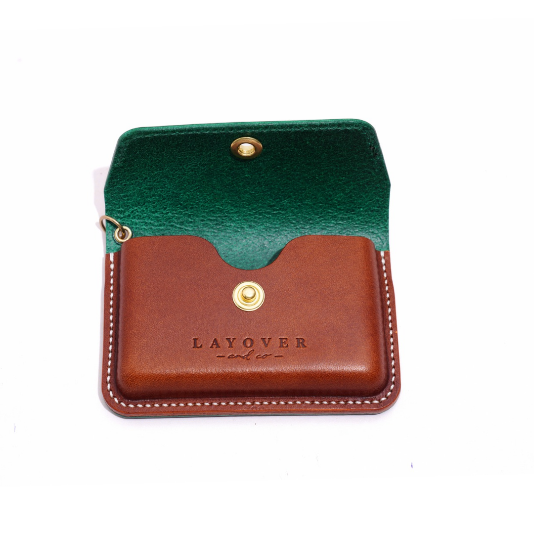 A close-up of an open handmade leather pouch featuring a rich brown body and forest green interior flap. The pouch has a brass snap button and detailed white stitching. The "Layover and Co." logo is embossed on the front.