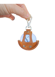 A close-up of a brown leather golf ball holder with visible stitching and metallic rivets, held by a hand. The holder features a secure clip at the top and an open-front design showcasing a white golf ball inside.