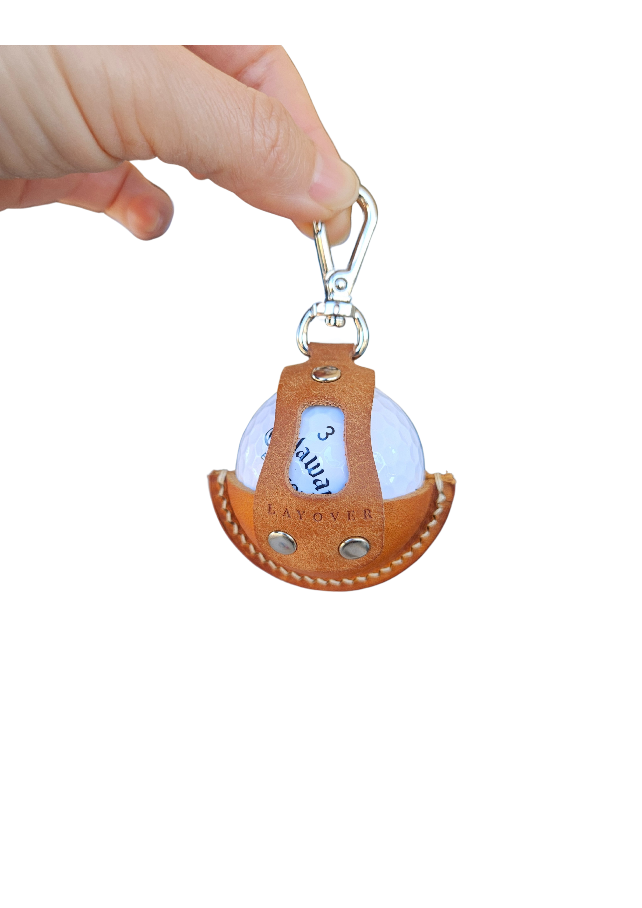 A close-up of a brown leather golf ball holder with visible stitching and metallic rivets, held by a hand. The holder features a secure clip at the top and an open-front design showcasing a white golf ball inside.
