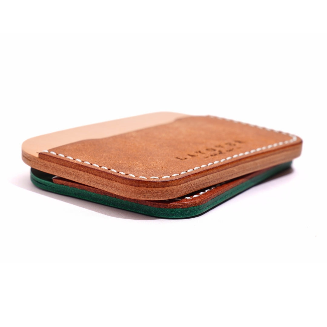 A slim, handcrafted leather card holder featuring a natural tan and brown exterior with white stitching and a green leather layer visible at the bottom. The minimalist design showcases Layover and Co.'s signature craftsmanship.