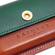 A detailed close-up of a handcrafted leather pouch from Layover and Co., featuring a green flap with matching stitching, a brass snap closure, and a rich brown base embossed with the brand’s logo.