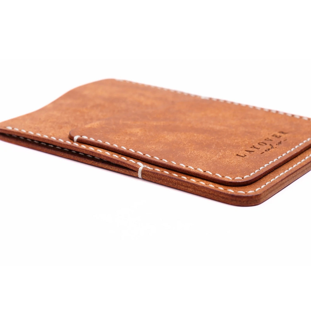 A slim, handcrafted leather wallet in a warm, rustic brown tone, featuring white hand-stitched detailing and a subtle embossed "Layover and Co." logo. The minimalist design includes a money clip and card slots for everyday use.